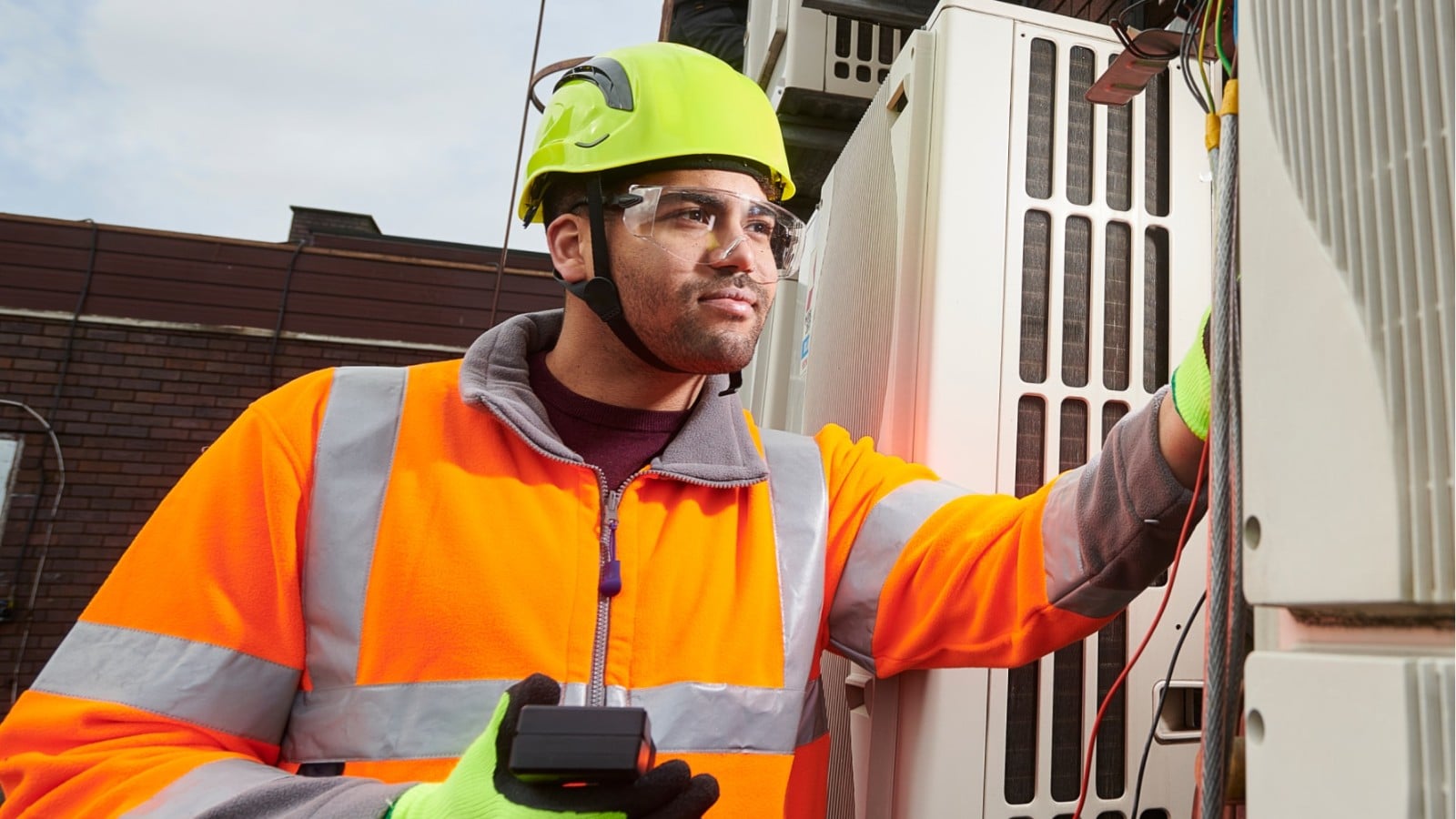 Technician in a hard hat, safety glasses and high-visibility jacket holding a handheld meter while checking rooftop HVAC units
