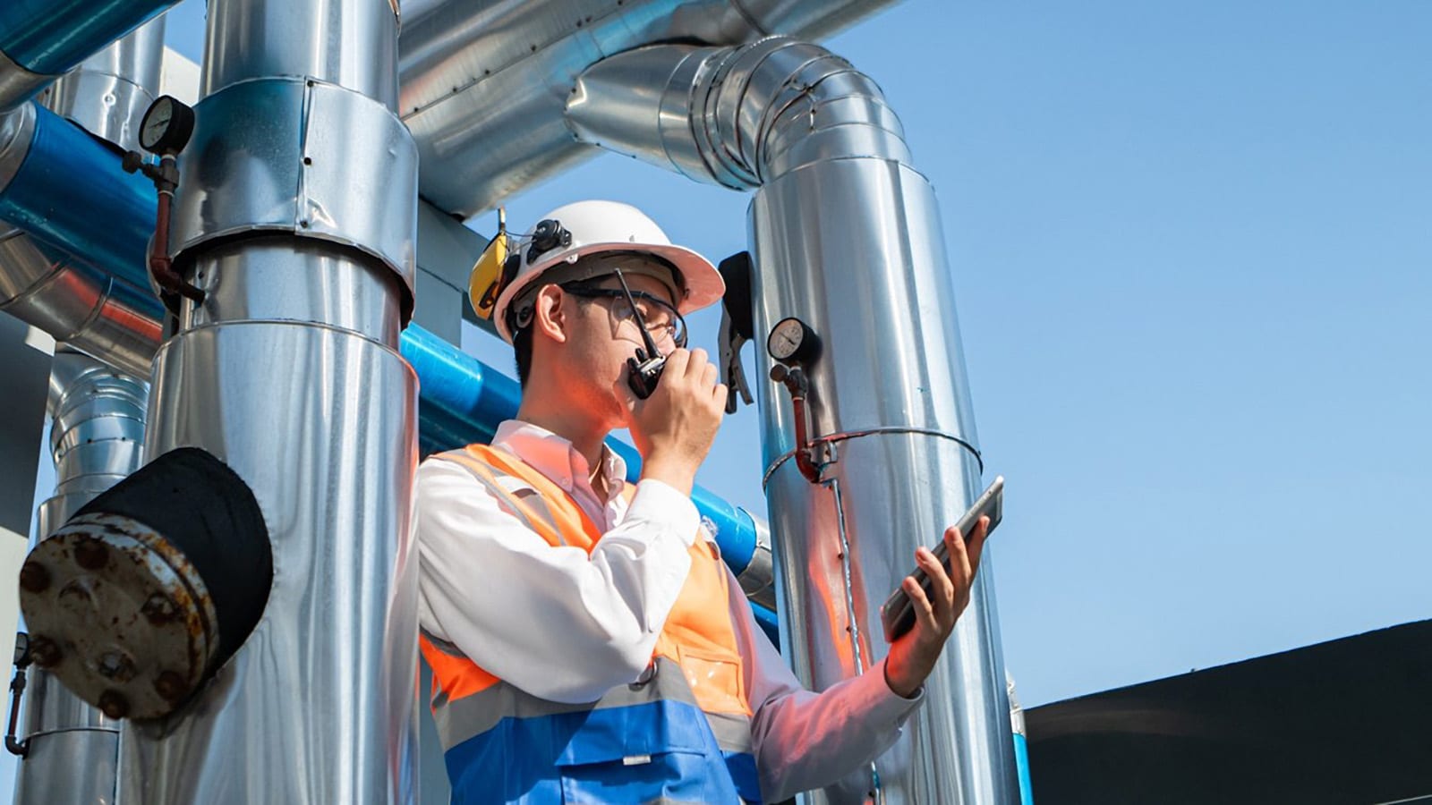 Engineer in a hard hat using a radio and tablet beside insulated mechanical piping